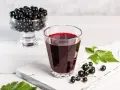 Szklanka z nalewką z czarnej porzeczki, obok kiść owoców i liście. W tle wazka pełna czarnych porzeczek.