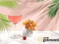 Szklanka z różowym napojem, kiść jasnych winogron i pojedyncze czerwone winogrono. W tle liście palmowe i różowa ściana.