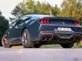 Ciemnoszary Ford Mustang, niczym wino dark horse, prezentuje swoje sportowe linie na tle zieleni. Widok z tyłu podkreśla mocne światła i podwójne końcówki wydechu.