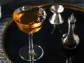 Elegancki koktajl w kolorze calvados, z wiśnią, obok kieliszka do mierzenia i buteleczki z bitters. Idealny do deserów.
