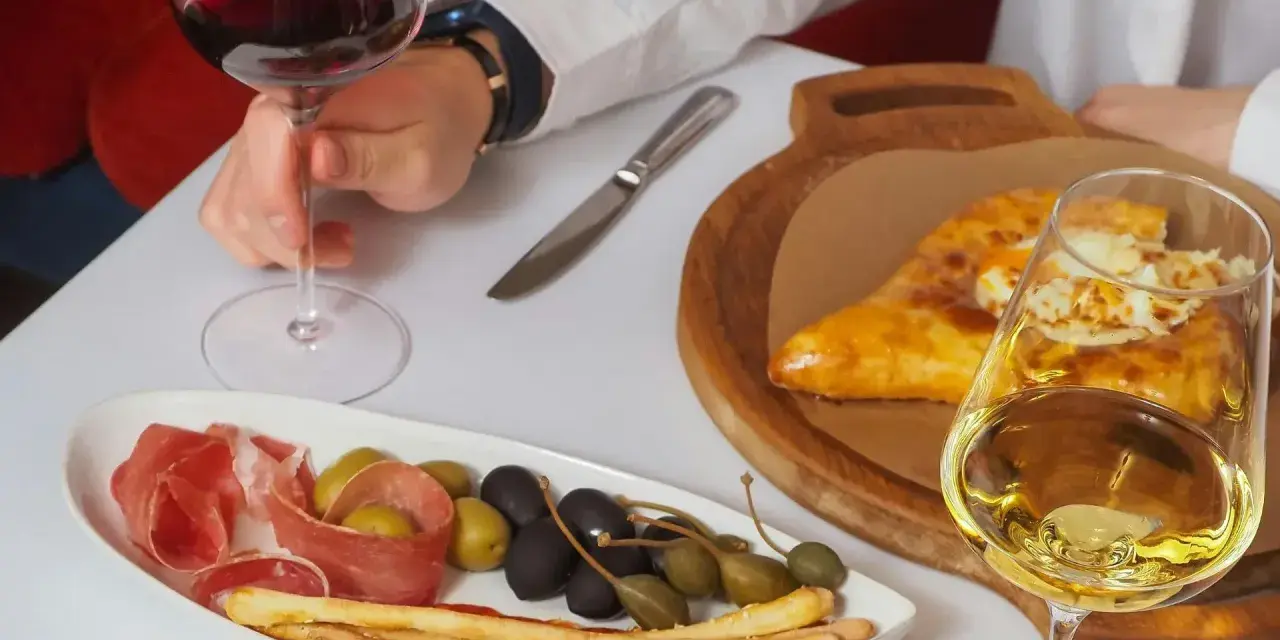 Ręka trzyma kieliszek czerwonego wina wytrawnego. Obok talerz z wędliną, oliwkami i grissini oraz chaczapuri i kieliszek białego wina.