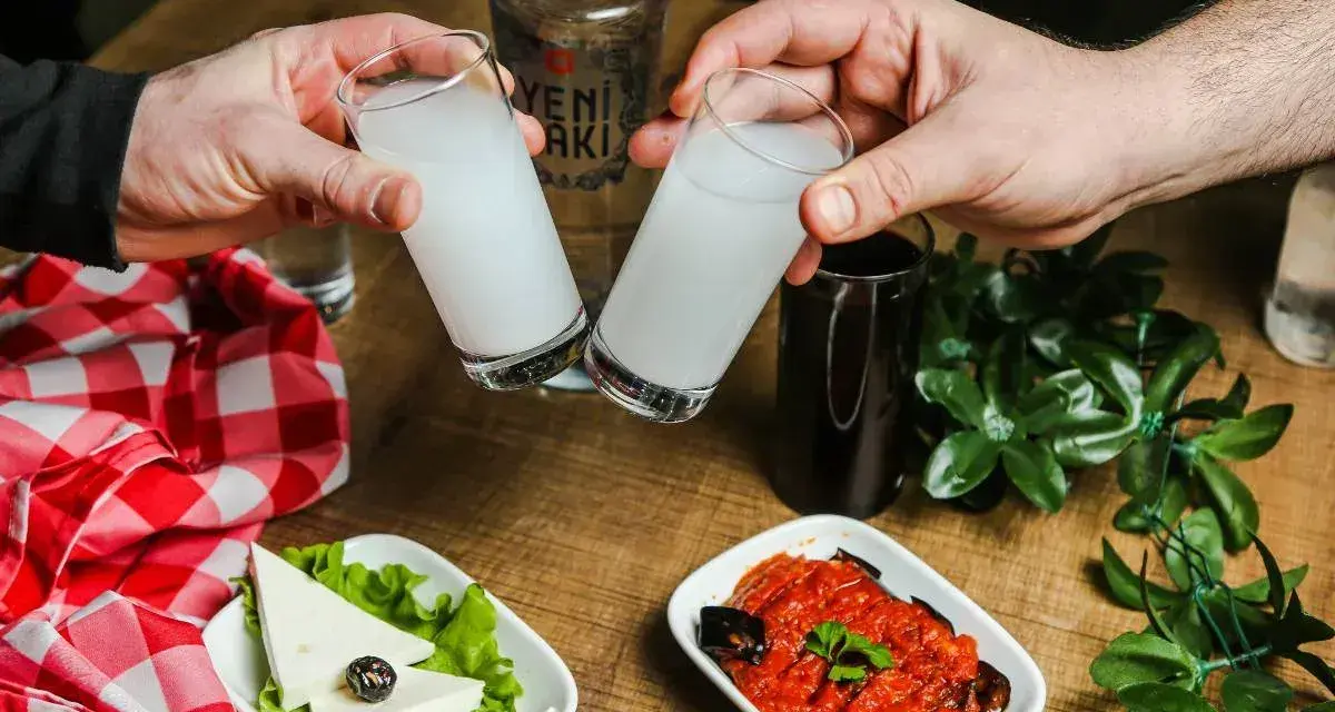 Dwie szklanki z rakı, tradycyjnym tureckim alkoholem, unoszą się do toastu. Obok ser, sałata, sos pomidorowy z bakłażanem i butelka rakı. Idealne połączenie na wieczór.