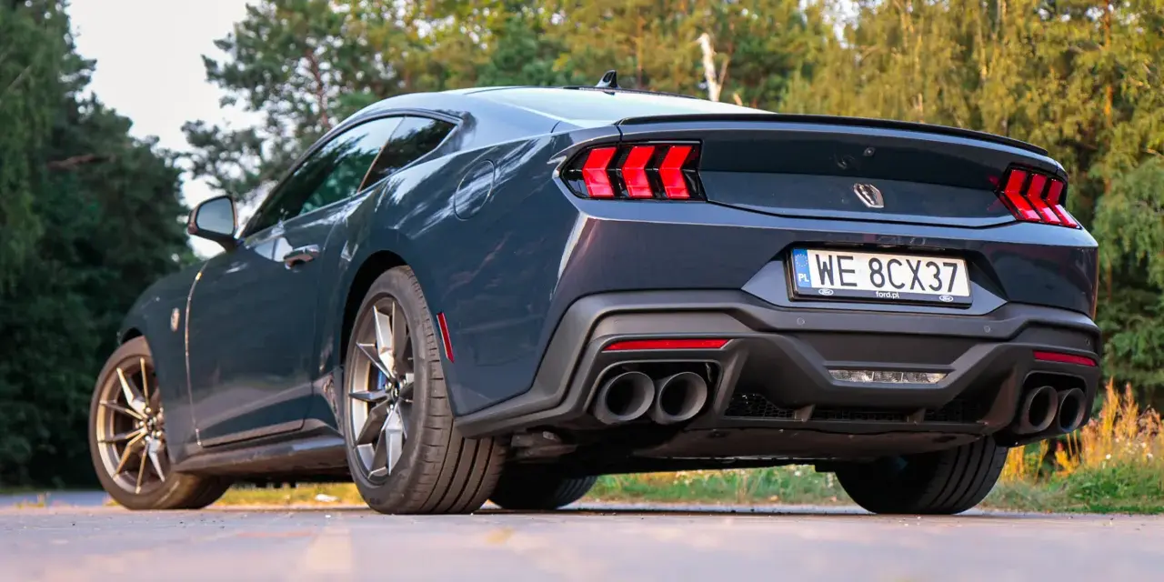 Ciemnoszary Ford Mustang, niczym wino dark horse, prezentuje swoje sportowe linie na tle zieleni. Widok z tyłu podkreśla mocne światła i podwójne końcówki wydechu.