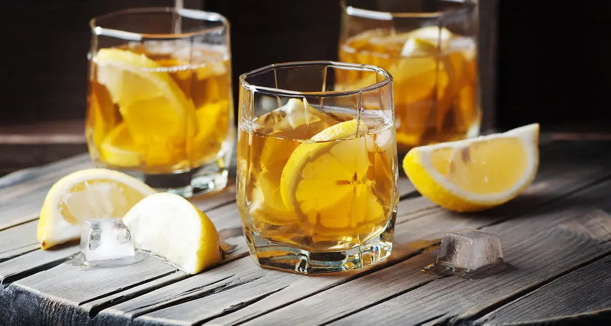 Metaxa orange z czym pić? Idealnie z lodem i plasterkiem cytryny. Trzy szklanki złocistego trunku na drewnianym blacie.