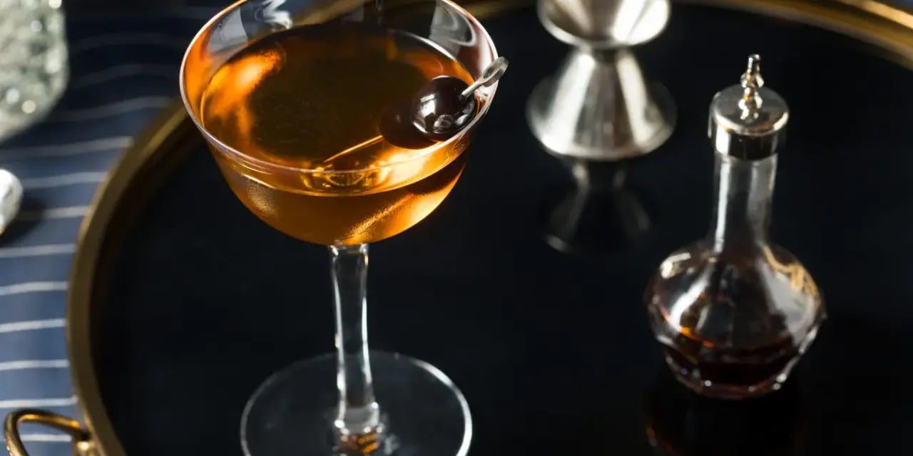 Elegancki koktajl w kolorze calvados, z wiśnią, obok kieliszka do mierzenia i buteleczki z bitters. Idealny do deserów.