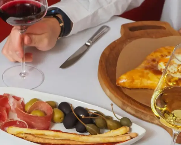 Ręka trzyma kieliszek czerwonego wina wytrawnego. Obok talerz z wędliną, oliwkami i grissini oraz chaczapuri i kieliszek białego wina.