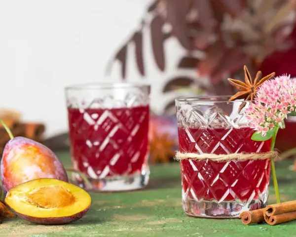 Dwa kieliszki z rubinową nalewką z śliwek, ozdobione kwiatami i anyżem. Obok świeże śliwki i laski cynamonu.