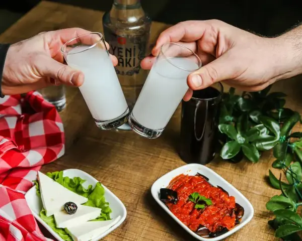 Dwie szklanki z rakı, tradycyjnym tureckim alkoholem, unoszą się do toastu. Obok ser, sałata, sos pomidorowy z bakłażanem i butelka rakı. Idealne połączenie na wieczór.