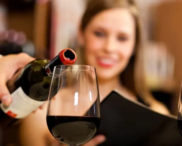 Kelner nalewa wino, a uśmiechnięta kobieta czeka na kieliszek. Wino zawiera siarczyny, które pomagają w jego konserwacji.