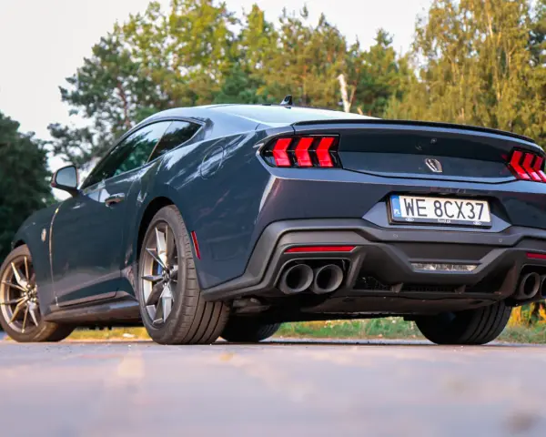 Ciemnoszary Ford Mustang, niczym wino dark horse, prezentuje swoje sportowe linie na tle zieleni. Widok z tyłu podkreśla mocne światła i podwójne końcówki wydechu.