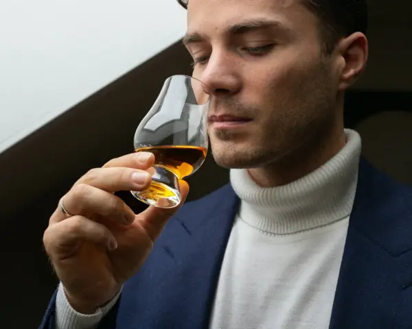 Mężczyzna w eleganckim garniturze i golfie wącha bursztynowy płyn w kieliszku. Czy to single malt?