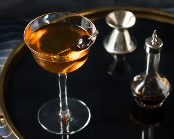Elegancki koktajl w kolorze calvados, z wiśnią, obok kieliszka do mierzenia i buteleczki z bitters. Idealny do deserów.
