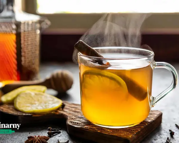 Rozgrzewająca herbata z whisky, z plastrem cytryny i laską cynamonu, paruje w szklance. Obok leżą cytryny i przyprawy.
