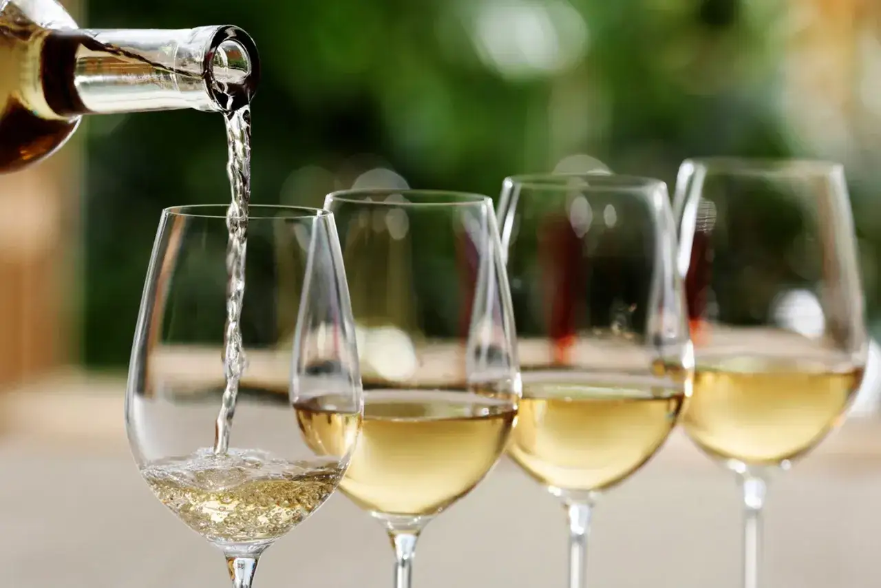 Białe wino, prawdopodobnie Pinot Grigio, nalewane do kieliszków. Wymowa nazwy wina jest kluczowa dla jego docenienia.