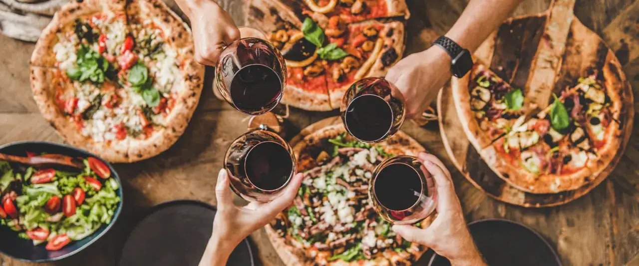 Ręce unoszą kieliszki z czerwonym winem do toastu nad pysznymi pizzami i sałatką. Idealne wino do pizzy na wspólny wieczór.