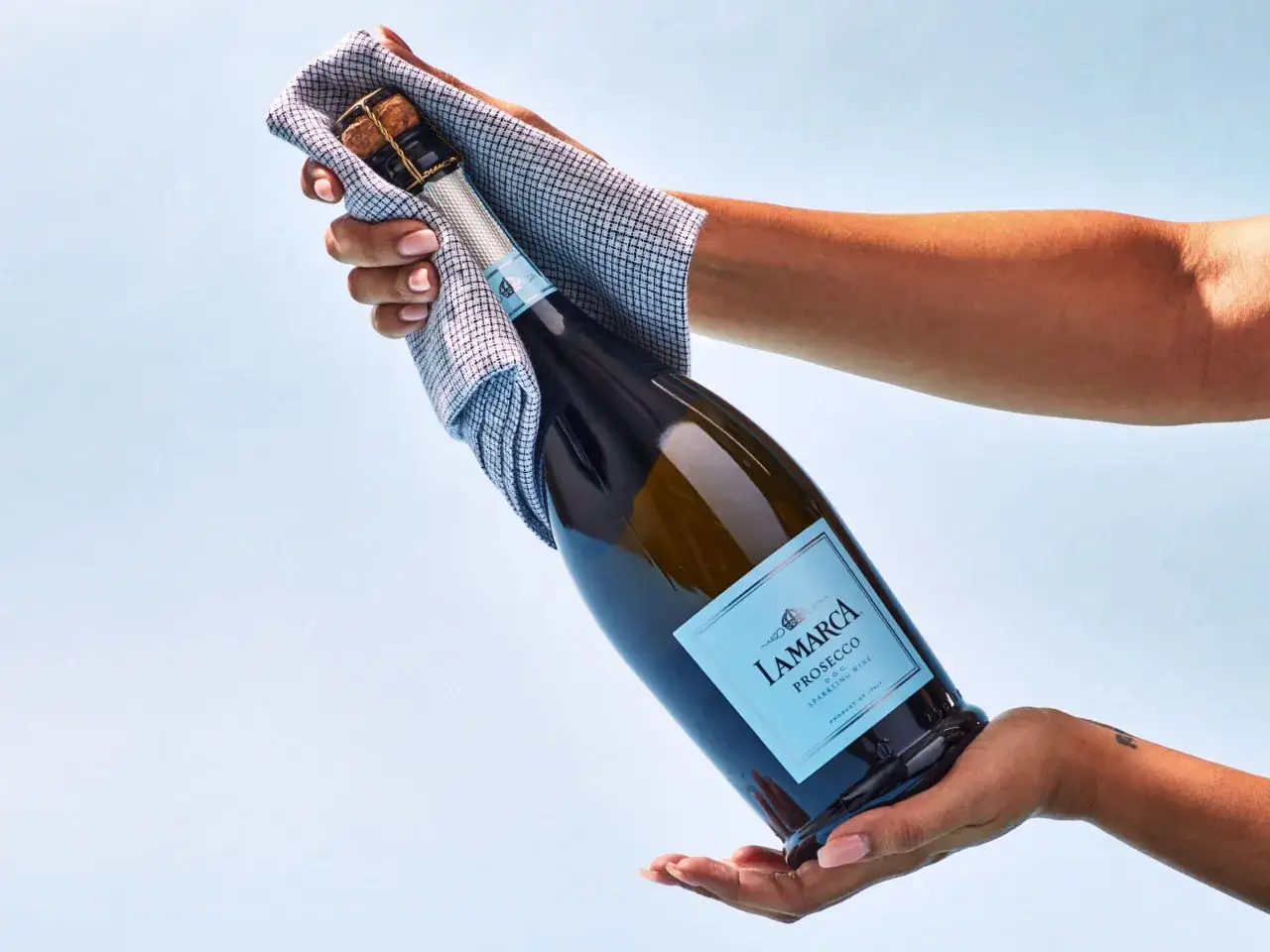 Dwie ręce trzymają butelkę prosecco, jedną owiniętą w ściereczkę. Oto jak otworzyć prosecco, by cieszyć się bąbelkami.