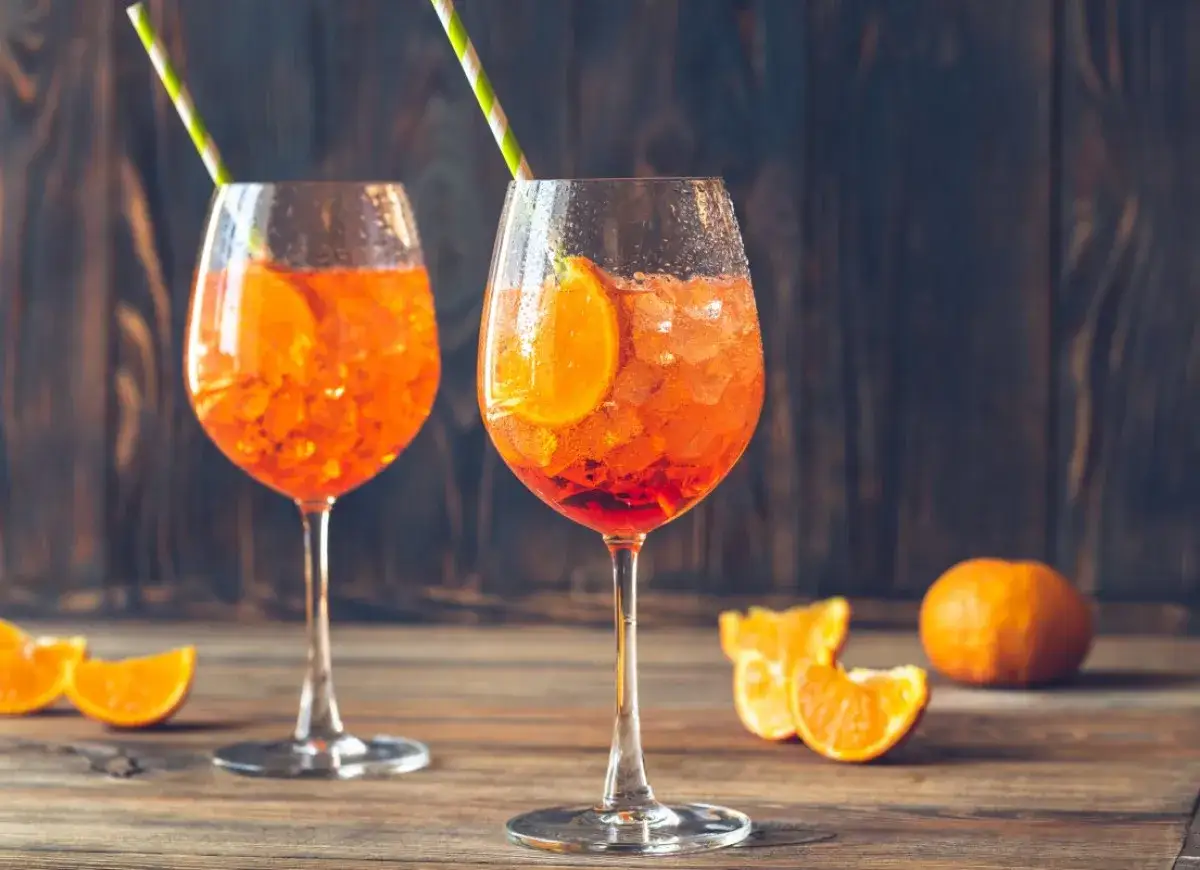 Dwa orzeźwiające Aperol Spritz z plasterkami pomarańczy i słomkami, na drewnianym stole.
