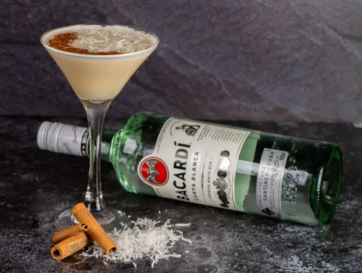 Koktajl z Bacardi z czymś kremowym, posypany wiórkami kokosowymi i cynamonem. Obok butelka Bacardi Carta Blanca, laski cynamonu i wiórki.