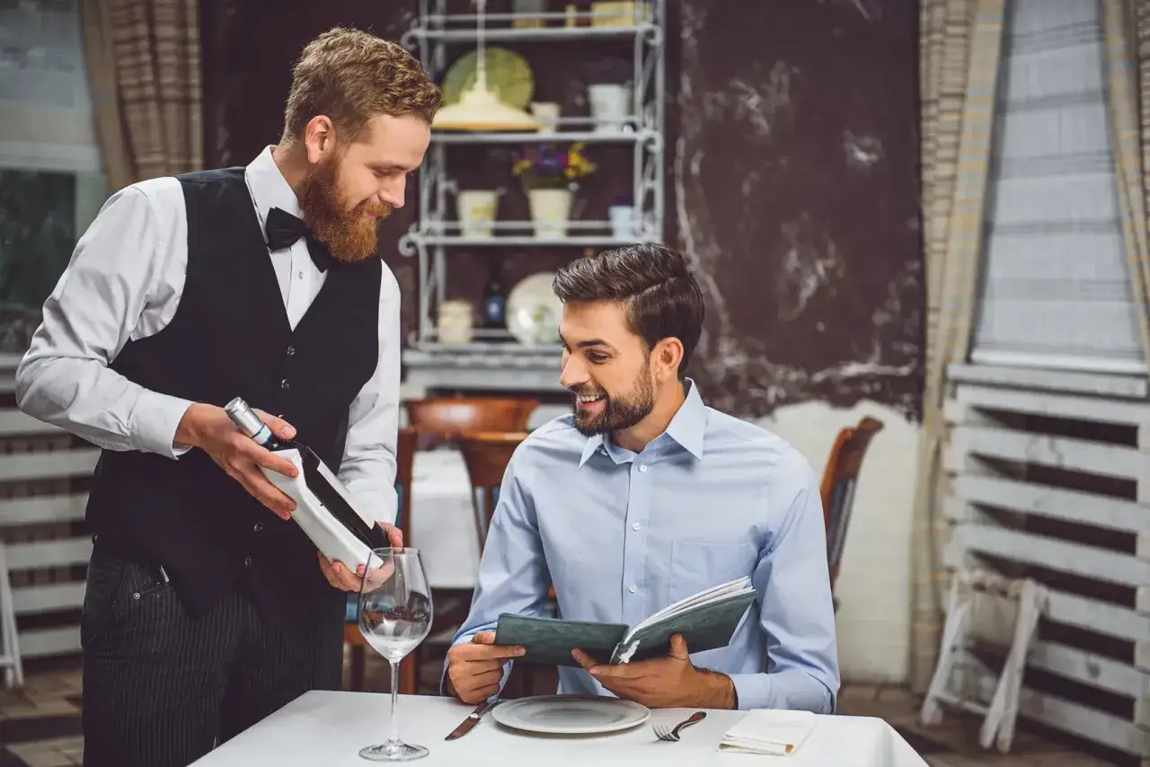 Sommelier, kto to? Mężczyzna w kamizelce prezentuje butelkę wina klientowi przeglądającemu kartę dań.