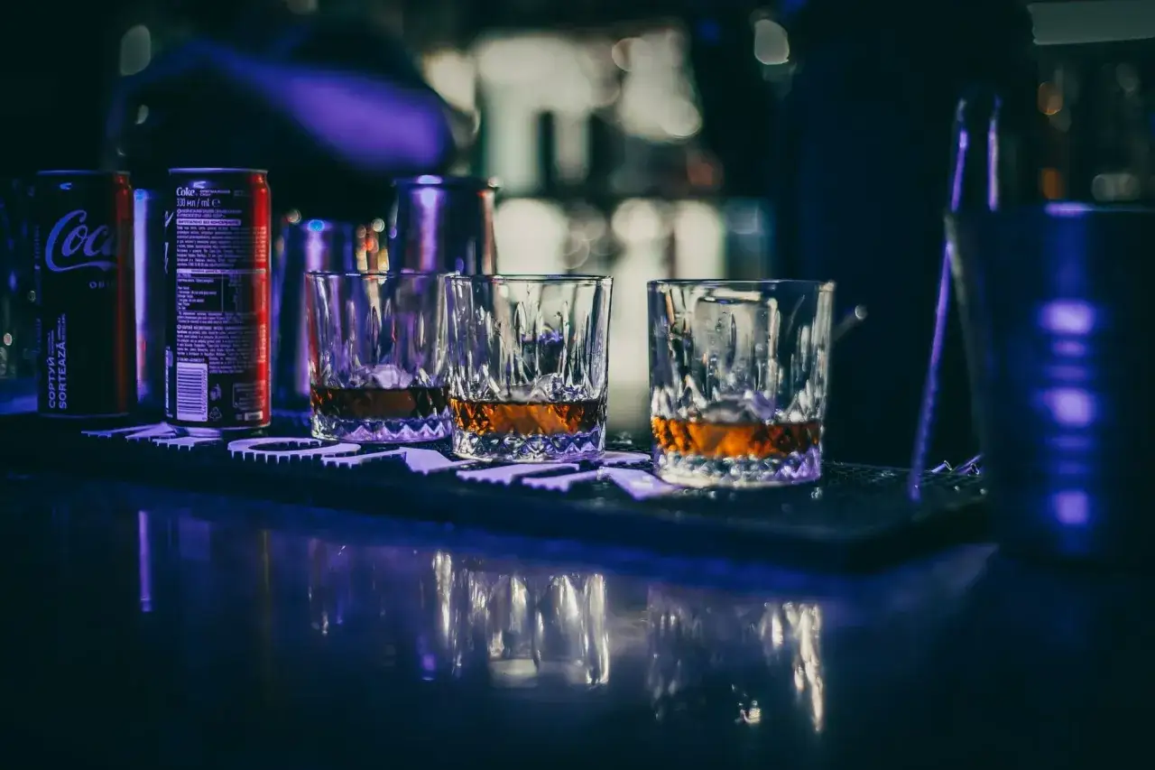 Trzy szklanki z bursztynowym płynem i lodem, obok puszki Coca-Coli. Idealna porcja whisky z lodem na barze.
