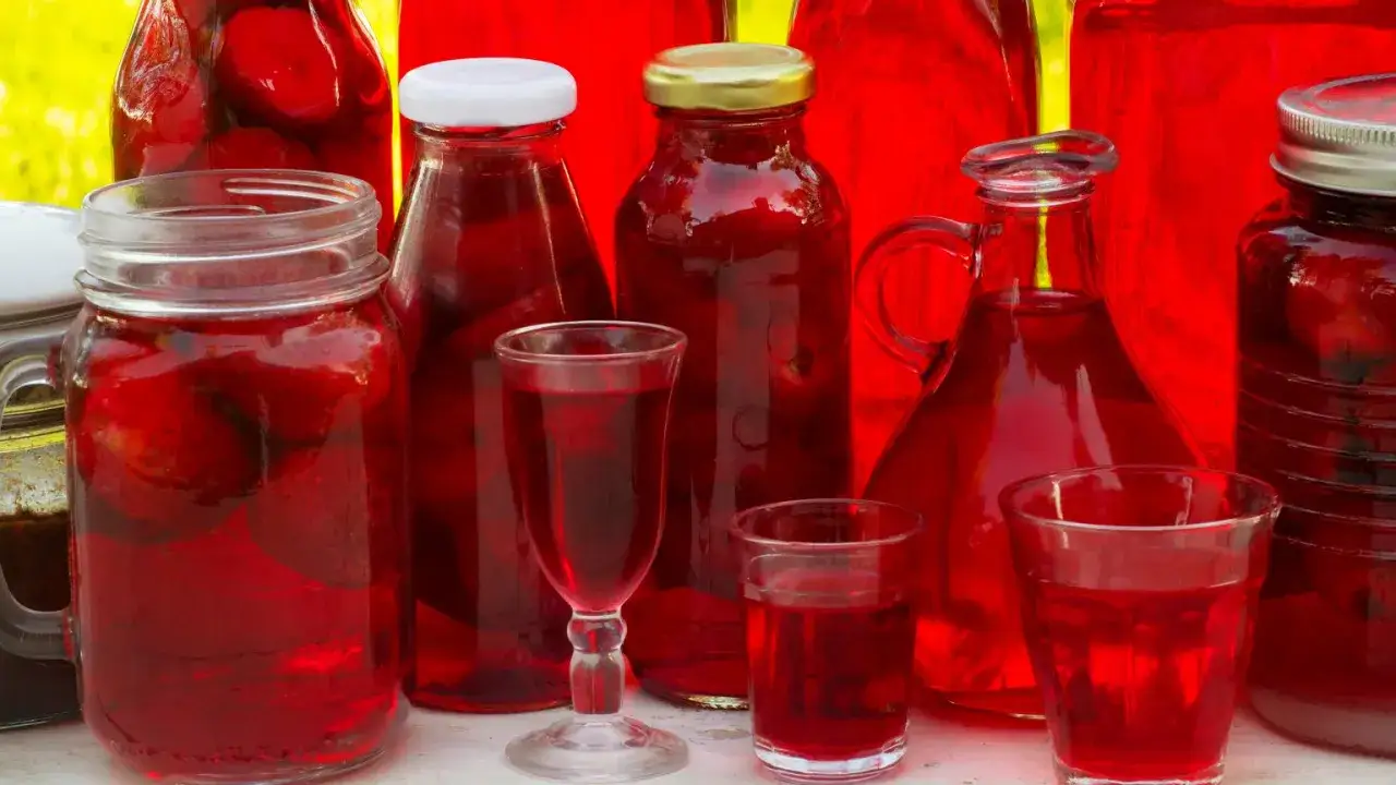 Słoje i butelki wypełnione rubinową nalewką z mrożonych truskawek, obok kieliszki z tym aromatycznym trunkiem.