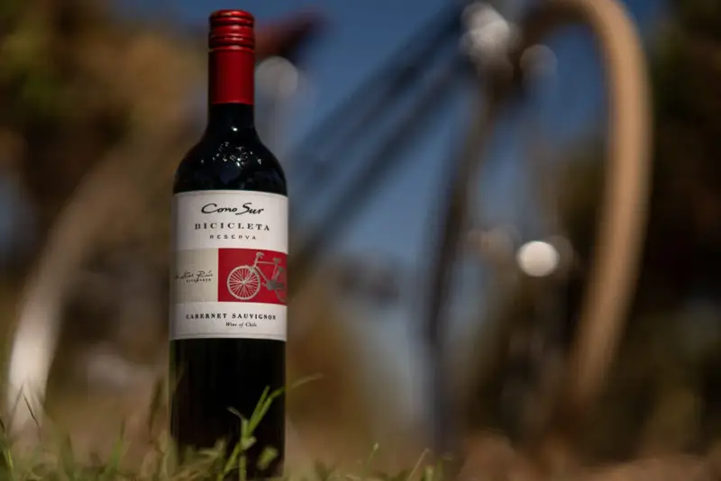 Butelka wina Cabernet Sauvignon z rowerem na etykiecie, stojąca na trawie.