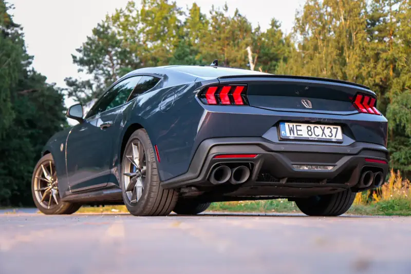 Ciemnoszary Ford Mustang, niczym wino dark horse, prezentuje swoje sportowe linie na tle zieleni. Widok z tyłu podkreśla mocne światła i podwójne końcówki wydechu.